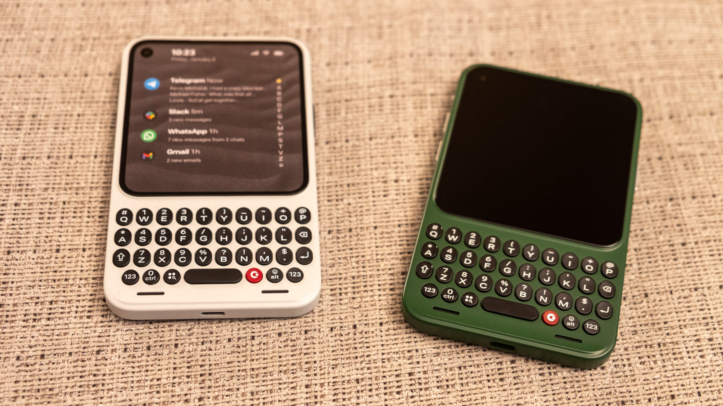 Two Clicks Communicator devices on a table, face up. The left one is white with the screen on showing some notifications and the right one is green with the screen off.